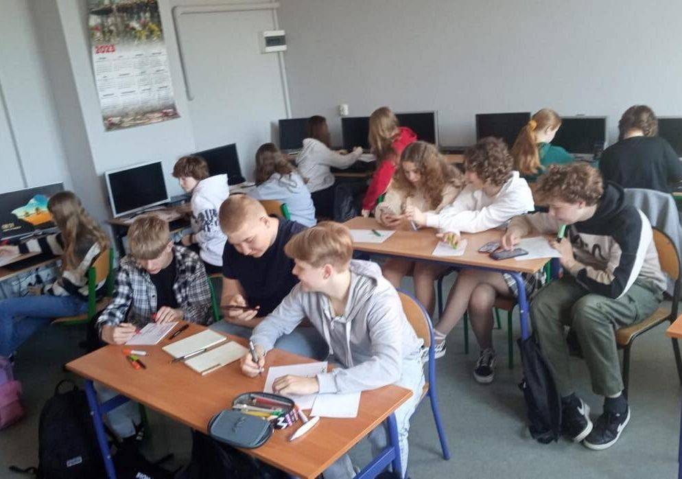 Uczniowie siedzą w ławkach szkolnych. Na stoikach leżą kartki i długopisy. Przy ścianie stoją komputery.