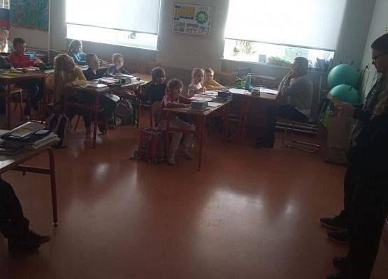 Uczniowie siedzą w ławkach szkolnych , odwróceni w stronę tablicy. Nauczyciel siedzi przy biurku przy oknie. Z przodu klasy stoi dziewczyna i chłopak.