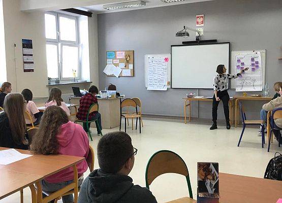 Klasa szkolna. Nauczyciel nakleja na tablice fioletowe karteczki i tłumaczy zadanie.