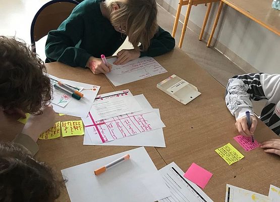 Uczennice siedzą w podwójnej ławce i piszą na kartkach kolorowymi mazakami