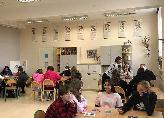 Klasa szkolna. Z przodu cztery uczennice siedzą w ławce, jedna ma ubraną koszule czerwoną w czarną kratę w okularach. Dwie dziewczynki mają różowe bluzki jedna czarną. Na ławkach leżą kartki i fotografie. Z tyłu klasy  z prawej strony 4 osobowa