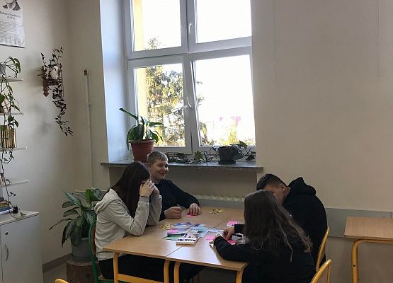 Klasa szkolna . Na wprost okno , przy którym stoją dwie połączone ławki szkolne. W ławkach siedzą na wprost siebie dwie uczennice i dwóch uczniów. Na ławkach leżą kartki i fotografie.