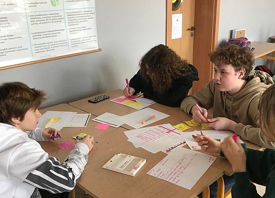 Uczennice siedzą w podwójnej ławce i piszą na kartkach kolorowymi mazakami