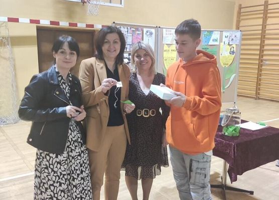 Uczeń w pomarańczowej bluzie , kobieta w beżowym komplecie ,dwie kobiety w sukienkach pozują do zdjęcia.