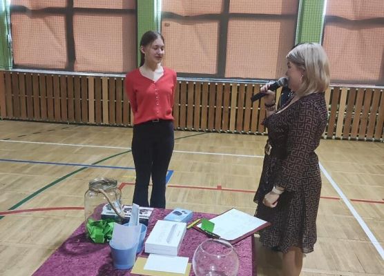 Kobieta mówi przez mikrofon do uczennicy. Druga kobieta wręcza nagrodę.