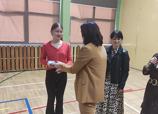 Kobieta mówi przez mikrofon do uczennicy. Druga kobieta wręcza nagrodę.