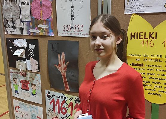 Uczennica trzyma w ręku nagrodę. Stoi przy tablicy z plakatami.