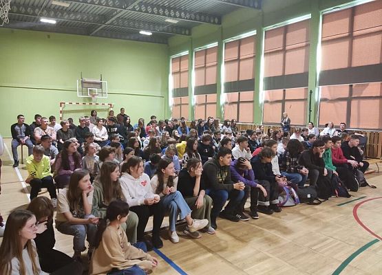 Uczniowie w dużej grupie siedzą na ławkach w sali gimnastycznej.