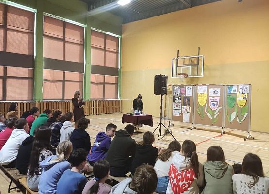 Grupa uczniów siedzi na ławce. Na przeciwko stoi stół z bordowym obrusem i czarny głośnik. Kobieta mówi przez mikrofon.