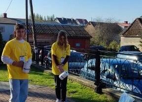 Z lewej strony stoi chłopak w żółtej koszulce trzyma w rękach puszkę, z prawej strony stoi dziewczyna w żółtej koszulce.