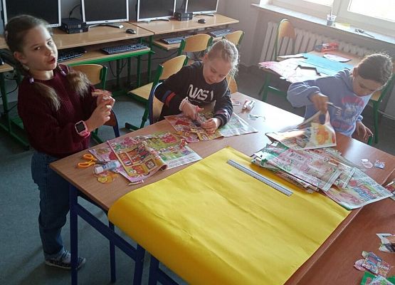 Na stole leży żółty brystol. Uczennice przyklejają kartki.