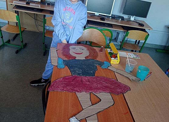 Na stoliku leży kartonowa postać dziewczynki. Uczennica stoi przy stoliku.