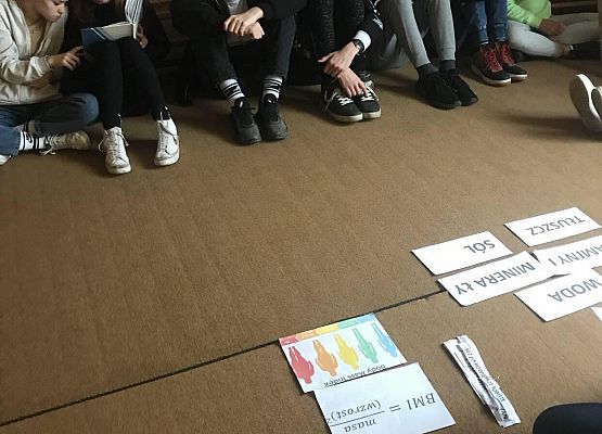 Uczniowie siedzą na dywanie. Na środku leżą materiały edukacyjne.