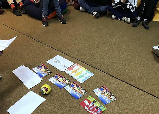 Uczniowe siedzą na dywanie. Na środku dywanu leżą materiały edukacyjne.