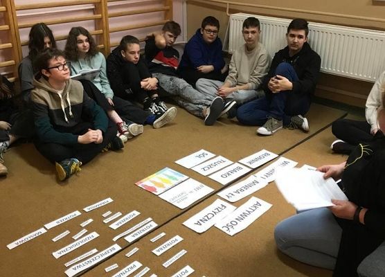 Grupa uczniow siedzi na dywanie. Na dywanie leżą kartki.