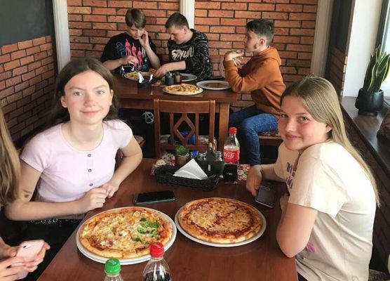 Uczniowie siedzą przy stolikach i jedzą pizzę.