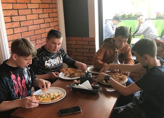 Uczniowie siedzą przy stolikach i jedzą pizzę.