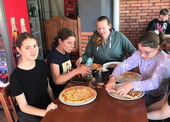 Uczniowie siedzą przy stolikach i jedzą pizzę.