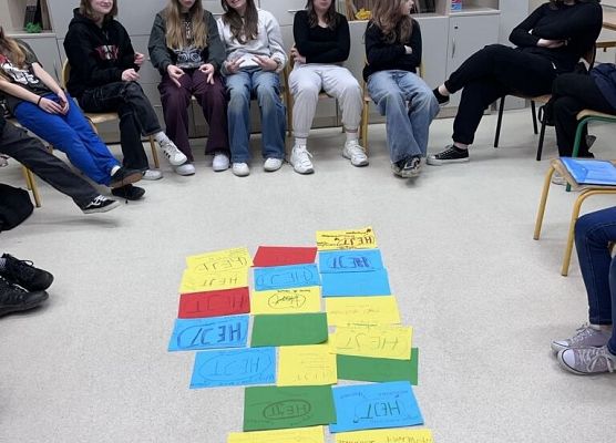 Na środku klasy na podłodze leżą kolorowe kartki a wokół nich siedzą uczniowie.