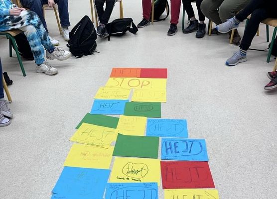 Na środku leżą na podłodze kolorowe kartki, wokół nich na krzesełkach siedzą uczniowie