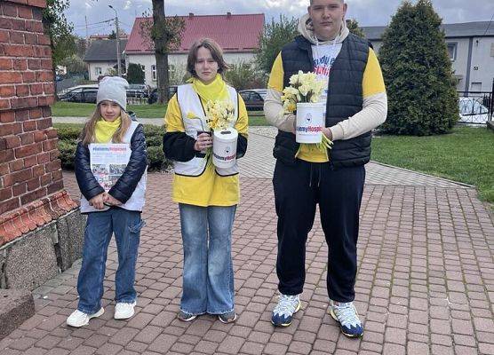 Uczniowie w żółtych koszulkach stoją przed kościołem. W rękach trzymają puszki na pieniądze i żonkile