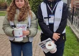 Uczniowie stoją przed kościołem. W rękach trzymają puszki na pieniądze i żonkile