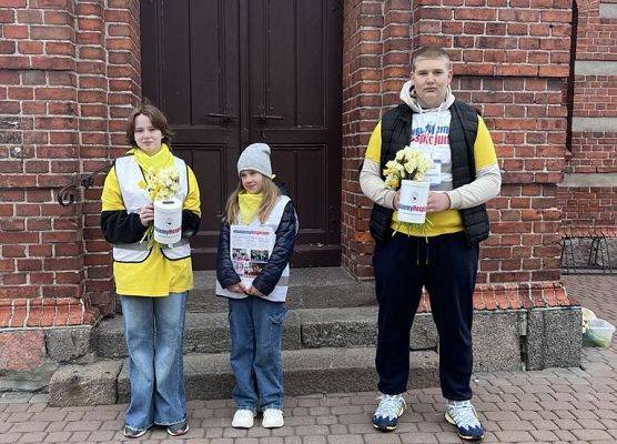Uczniowie w żółtych koszulkach stoją przed kościołem. W rękach trzymają puszki na pieniądze i żonkile