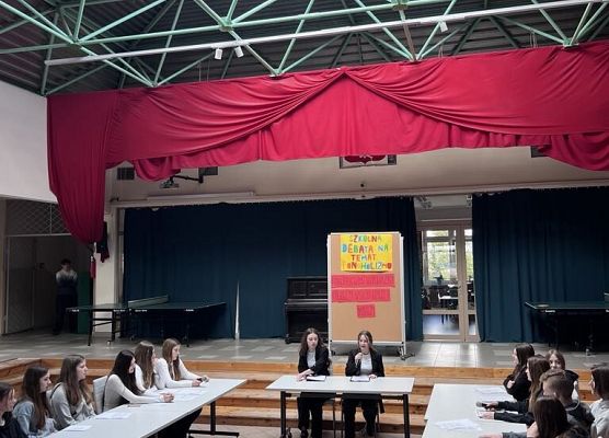Na środku patio stoją stoły, przy których siedzą uczniowie, trzymają mikrofon w rękach. Z tyłu stoi tablica z hasłem Debata
