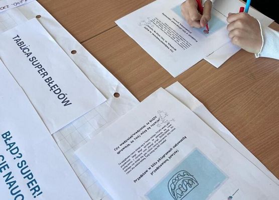 Na stole leżą kartki z zadaniami dla uczniów. Uczniowie wypełniają je mazakami