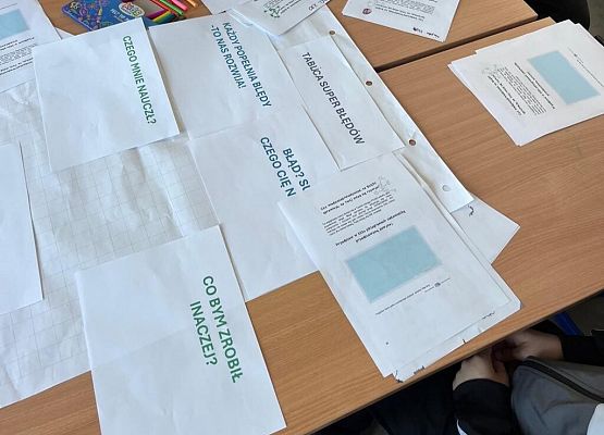 Na stole leżą kartki z zadaniami dla uczniów. Uczniowie wypełniają je mazakami