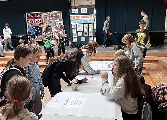 Uczniowie siedzą i stoją przy stoliku na którym są duże białe arkusze papieru