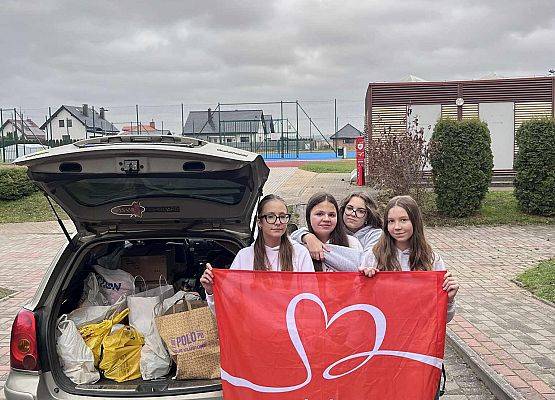 Uczniowie trzymają plakat czerwony z napisem Szlachetna paczka obok samochodu